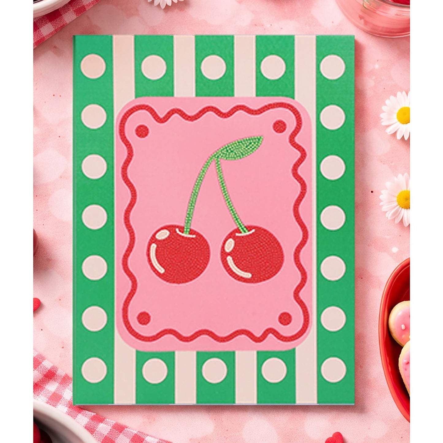 A square decorative item featuring a pink center with two red cherries and a green stem, surrounded by a green-and-white polka dot border on a pink background.