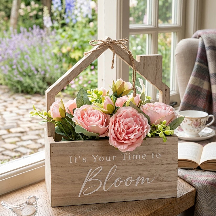 A wooden house-shaped floral planter box with pink roses and green leaves, sitting on a wooden windowsill in a cozy room setting with a window view of a garden, a teacup, and an open book.