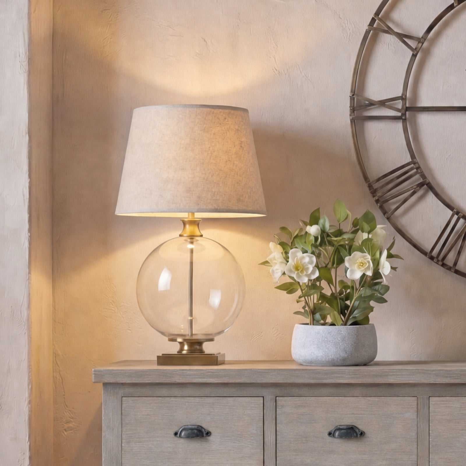 Table lamp with a glass shade and a potted plant on a wooden surface, with a clock in the background.