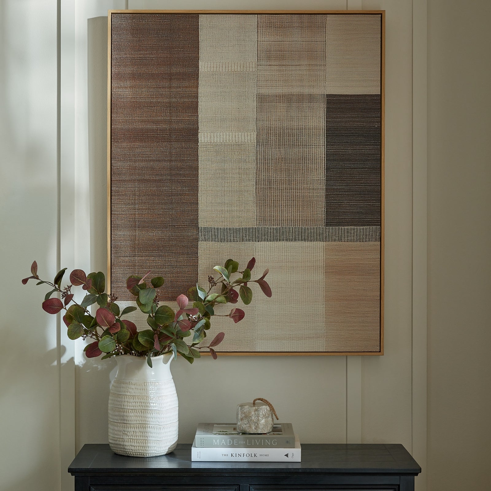 Framed abstract art on a wall above a table with a vase, books, and a small sculpture.