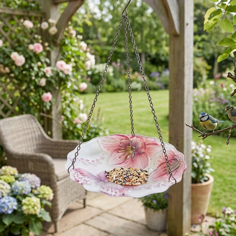 A hanging bird feeder with a pink floral plate design, filled with birdseed, suspended from a wooden pergola in a garden setting with a wicker chair and greenery in the background.