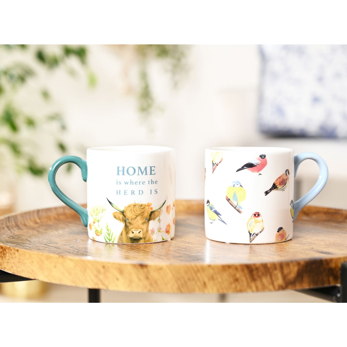 Two mugs on a wooden table with one featuring a cow and the other with bird illustrations.