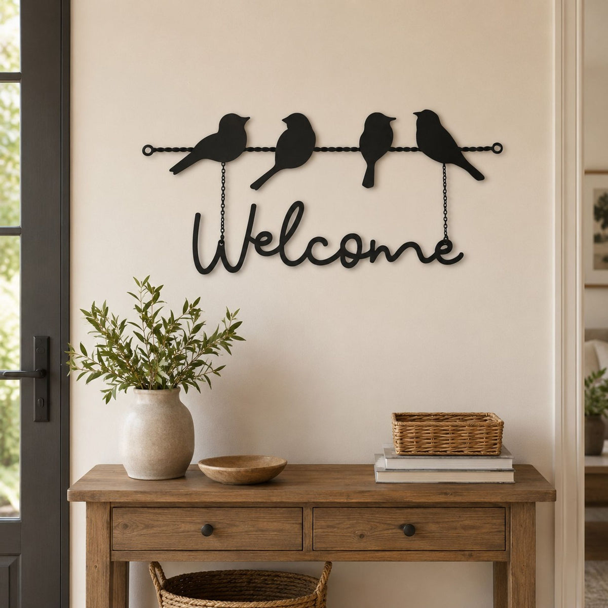 Black metal wall decor featuring three birds perched on a wire above the word 'Welcome', displayed on a beige wall above a wooden console table with a vase and basket.