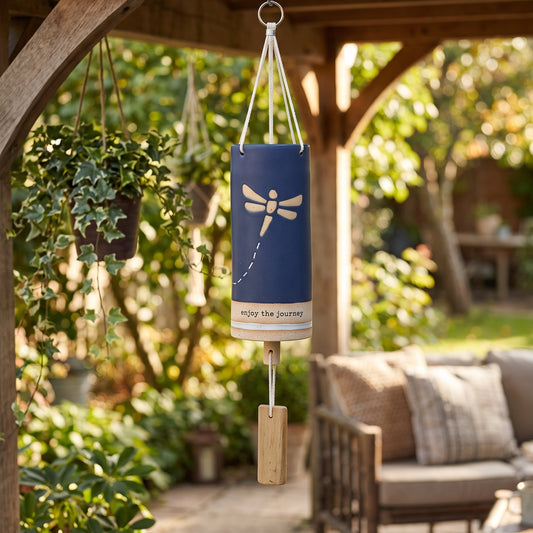 A hanging wind chime with a navy blue cylindrical body, a white dragonfly design, and the words 'Enjoy the Journey' on a wooden post in a garden patio setting.