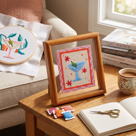 A framed embroidered cocktail-themed artwork stands on a wooden table beside an open book, scissors, thread spools, a mug, and stacked books in a bright living-room setting. Another embroidery hoop with a similar design rests on a chair in the background.
