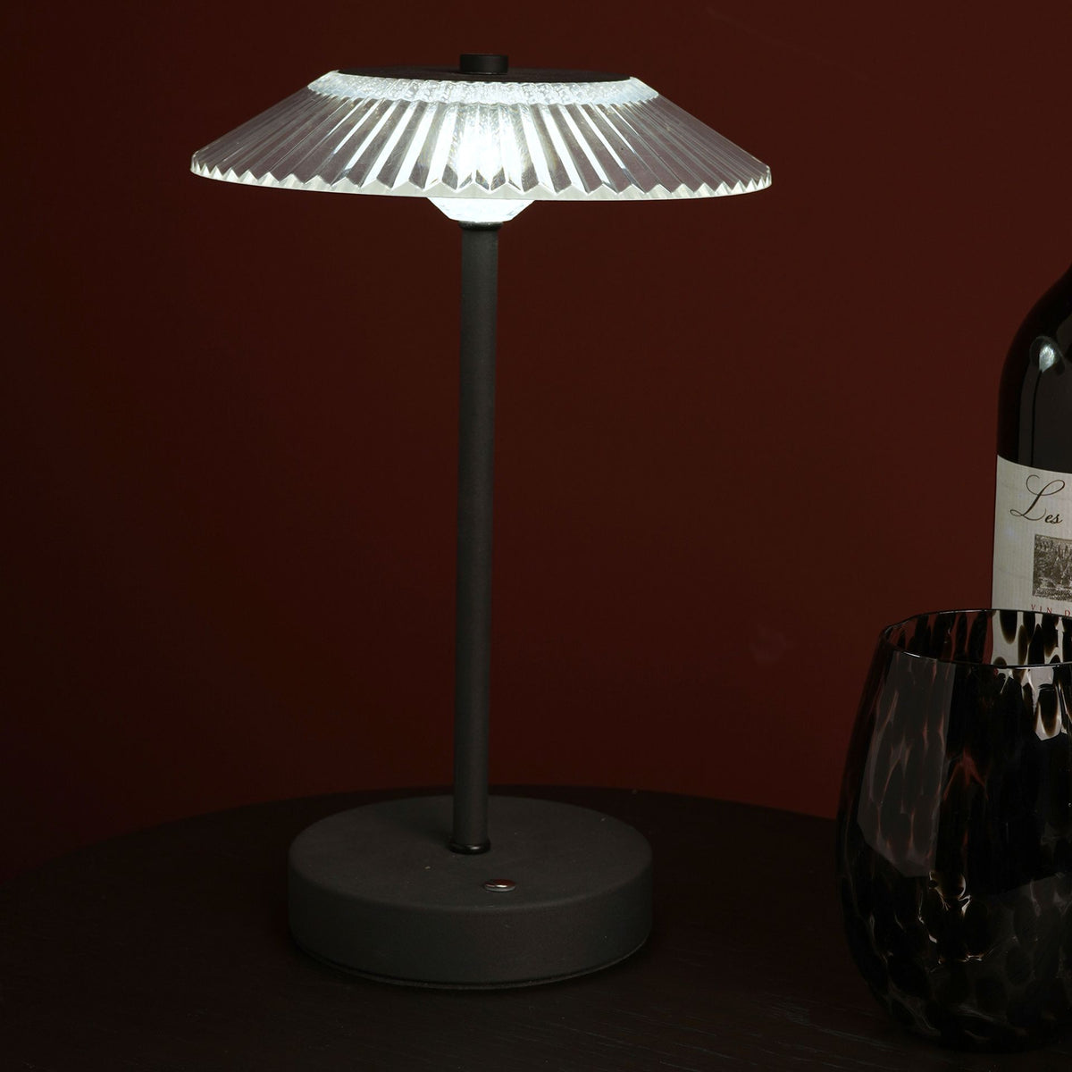 Table lamp with a dark base and white shade next to a bottle on a dark surface.