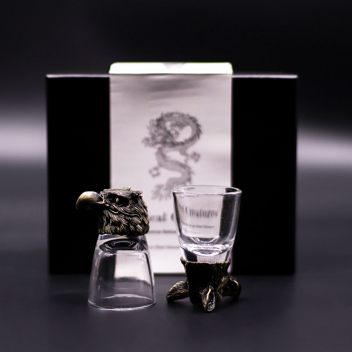 Two shot glasses with decorative stands in front of a branded box on a dark background