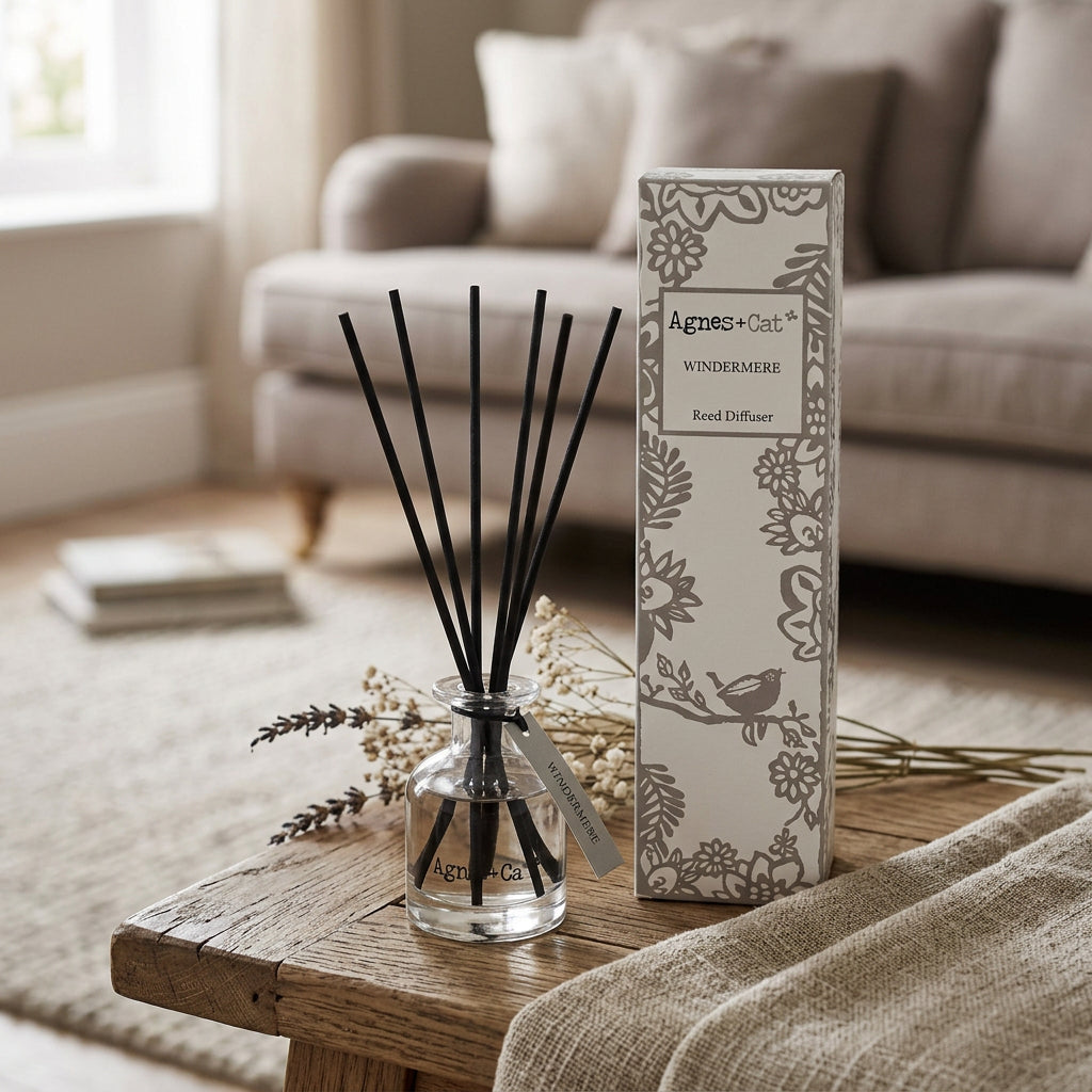 A clear glass reed diffuser bottle with black reeds and a hanging paper tag, shown next to its white floral-patterned box labeled Agnes & Cat Windermere Reed Diffuser on a white background.